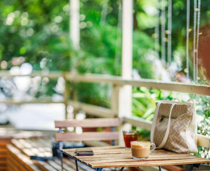 A stylish anti-theft crossbody bag with secure zippers is attached to a chair in a bustling cafe, with a coffee cup and travel guide visible.