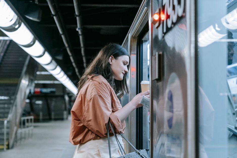 A busy professional woman quickly reapplying sunscreen mist to her face while on a public transport commute, with city views outside the window.