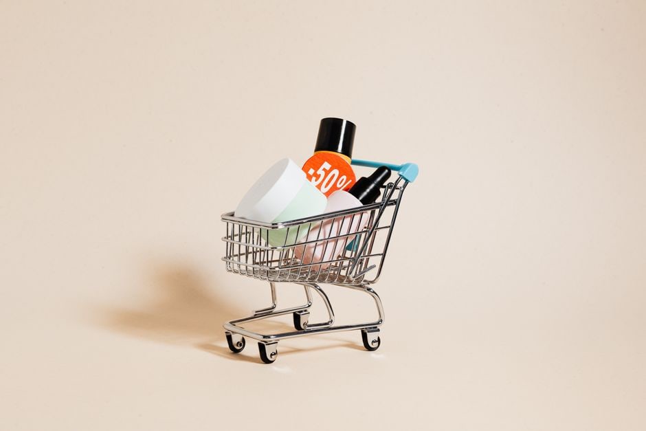 A grocery shopping cart filled with fresh produce and household items, with a hand holding a paper coupon over it, symbolizing smart shopping.