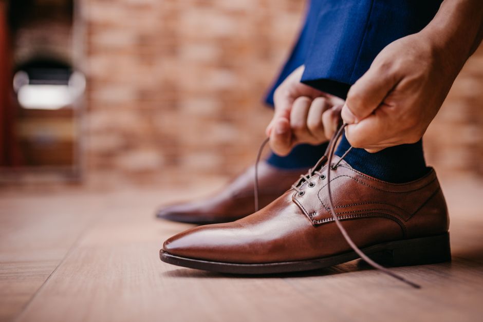 A close-up shot of a man's hands tying the laces of polished brown leather dress shoes, emphasizing the importance of well-maintained footwear for a sophisticated look.