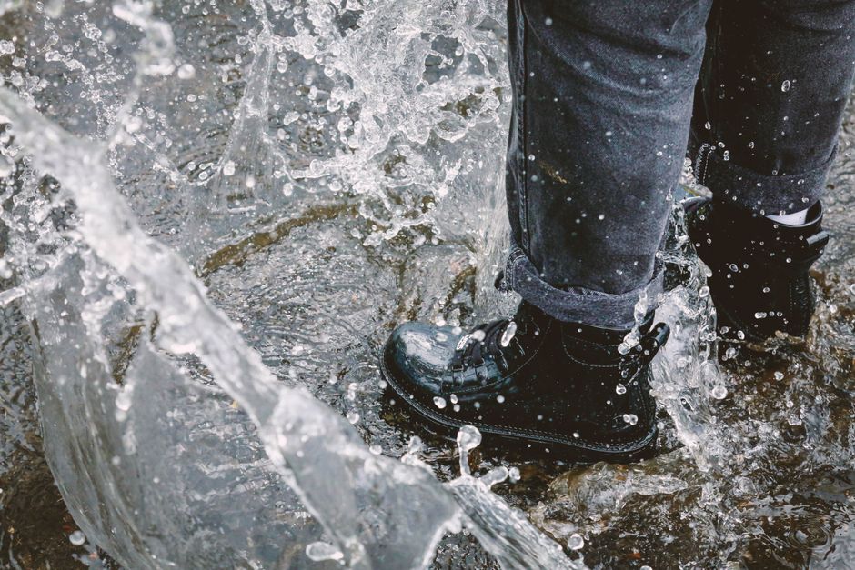 Close-up of a commuter's feet, clad in stylish waterproof boots, stepping confidently over a large puddle on a rainy city sidewalk.