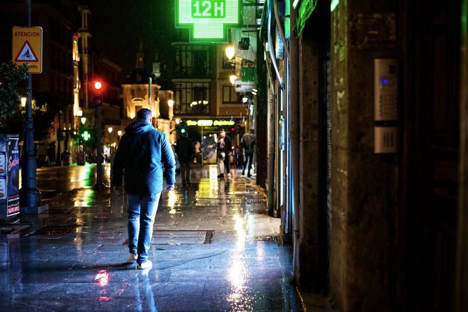 A commuter in a trench coat and waterproof boots hurrying across a crosswalk during a heavy rain shower in a European city.