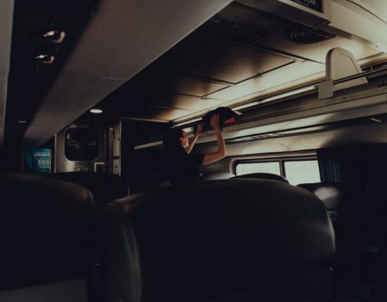 A sleek, small black travel bag with multiple compartments sitting on a train seat next to a window, suggesting a journey through an urban landscape.