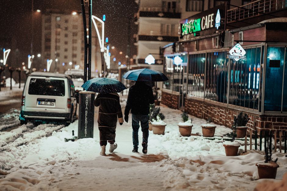 Two people bundled in winter coats and comfortable boots walking through a snowy, tree-lined street in a European city.