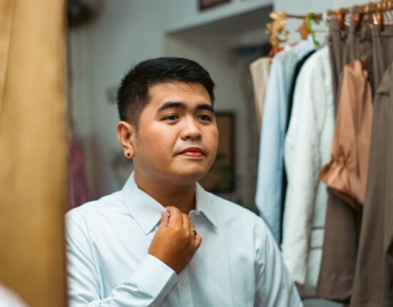 A man in a well-fitted light blue button-down shirt standing in front of a mirror, adjusting his collar, preparing for a first date or dinner.