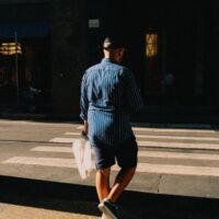 A man dressed in a light blue linen shirt and tailored beige shorts walking confidently down a sunny European city street, embodying practical men's summer fashion.