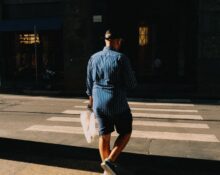 A man dressed in a light blue linen shirt and tailored beige shorts walking confidently down a sunny European city street, embodying practical men's summer fashion.