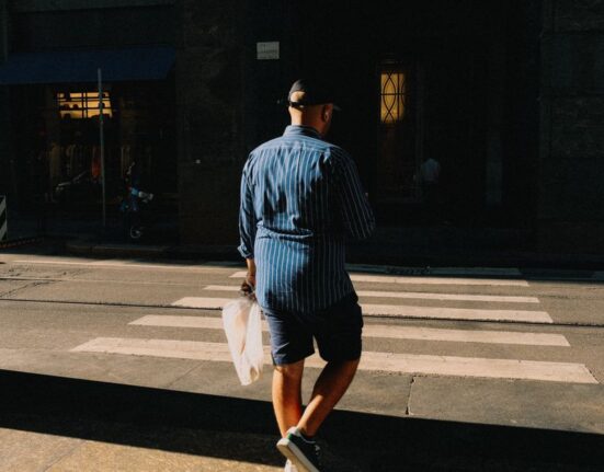 A man dressed in a light blue linen shirt and tailored beige shorts walking confidently down a sunny European city street, embodying practical men's summer fashion.
