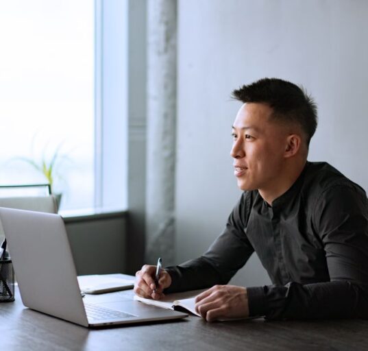 A man in smart casual attire, wearing a light blazer and dark trousers, sitting comfortably at a modern desk with a laptop, reflecting the hybrid work environment.