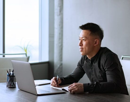 A man in smart casual attire, wearing a light blazer and dark trousers, sitting comfortably at a modern desk with a laptop, reflecting the hybrid work environment.
