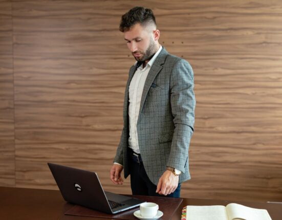 Man in navy blazer and light chinos working on a laptop in a modern, bright office setting.