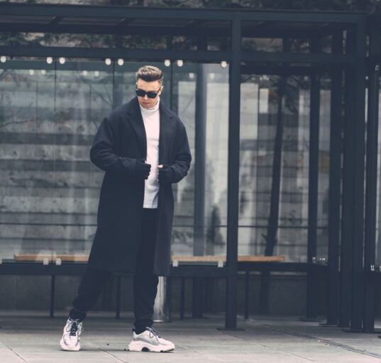 A man in a city setting, demonstrating effective winter layering with a wool overcoat, scarf, and gloves.