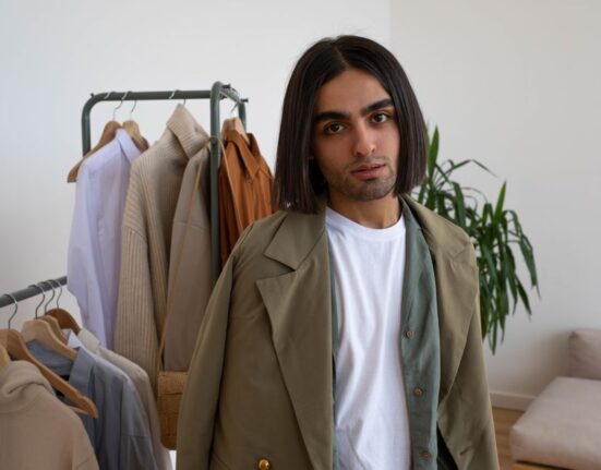 A man in a clean, organized bedroom looks thoughtfully at a minimalist closet filled with a few carefully chosen clothes on hangers.