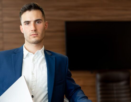 A man in a navy unstructured blazer and khaki chinos working on a laptop in a modern, well-lit office.