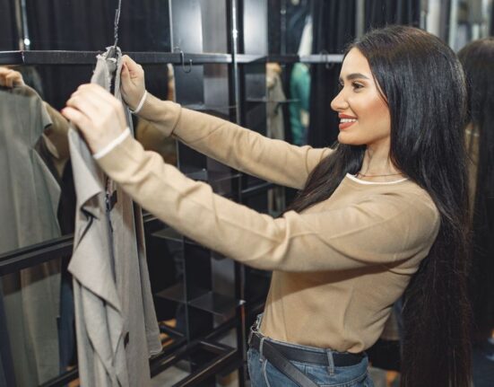 A man in a bright, organized room standing in front of a full-length mirror, adjusting the fit of a new pair of blue jeans, checking the length and waist.