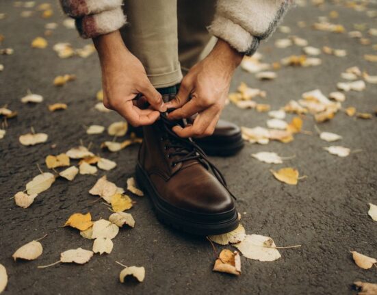 A man in tailored trousers sitting down, adjusting the laces of a pair of comfortable, dark brown leather derby shoes.
