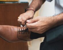 A man in a navy suit sitting down, adjusting the laces of his brown leather derby shoes, symbolizing the start of a comfortable workday.