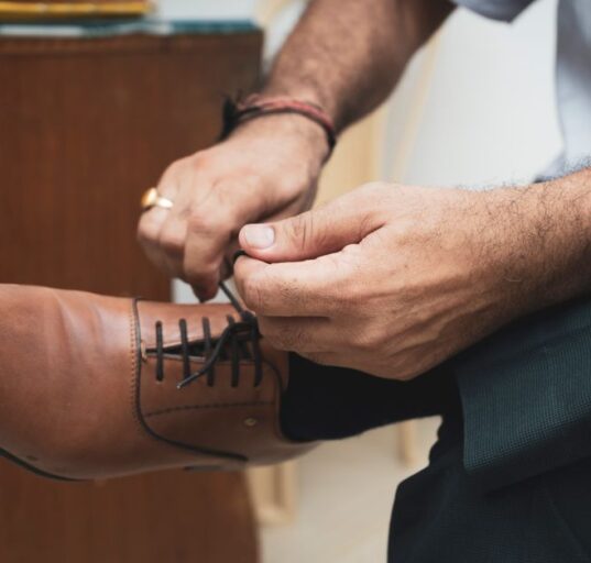A man in a navy suit sitting down, adjusting the laces of his brown leather derby shoes, symbolizing the start of a comfortable workday.