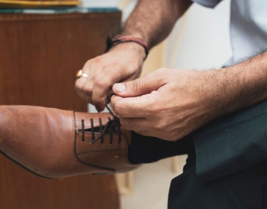A man in a navy suit sitting down, adjusting the laces of his brown leather derby shoes, symbolizing the start of a comfortable workday.