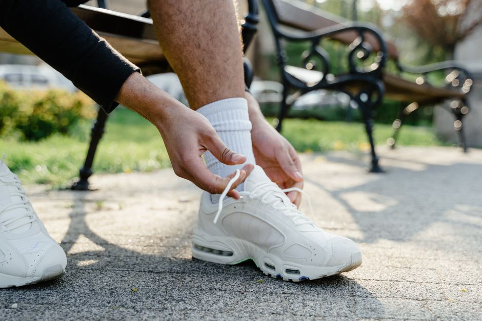 A man in casual attire is bending down to tie the laces of his budget-friendly walking shoes in a green urban park setting.
