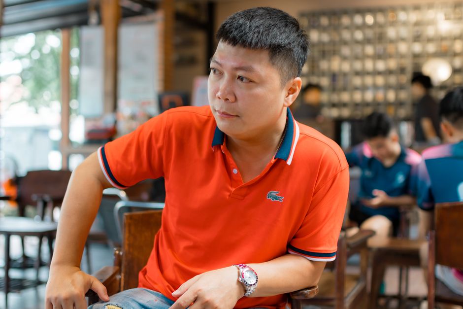A man seated at an outdoor cafe, wearing a light grey unstructured blazer over a dark polo shirt, paired with sunglasses, showcasing a smart casual summer look for professional settings or social events.