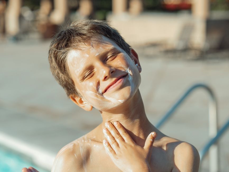 A person with clear, healthy skin smiling while gently applying a broad-spectrum SPF sunscreen to their face outdoors, highlighting daily sun protection.