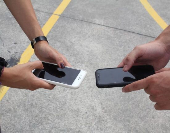 A person holding a smartphone, comparing prices on a screen while looking at a laptop with shopping websites open.