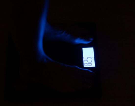 A person sitting on the floor, measuring their foot length with a tape measure against a wall, with a piece of paper and pencil nearby.