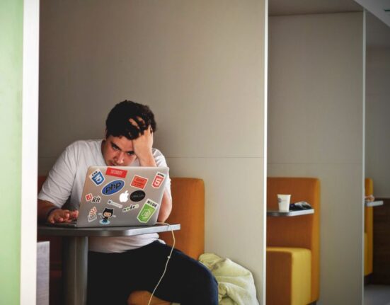 Woman looking stressed while shopping on a laptop, surrounded by gift bags, symbolizing last-minute pressure.