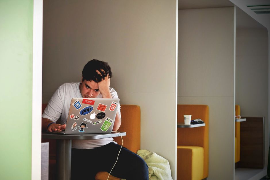 A person with a worried expression, sitting at a laptop, surrounded by gift boxes and shopping bags, clearly stressed about last-minute online gift shopping.