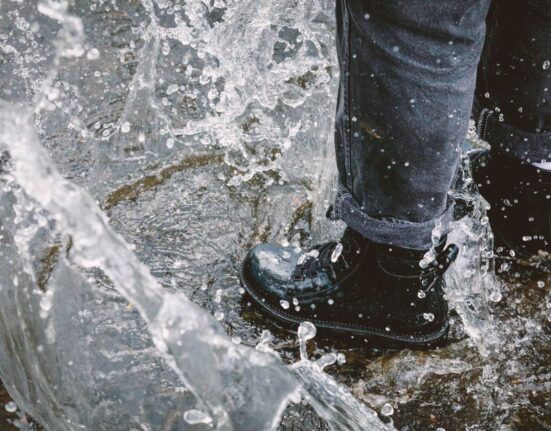 A person wearing dark waterproof shoes walking quickly across a rain-slicked city pavement with reflections.