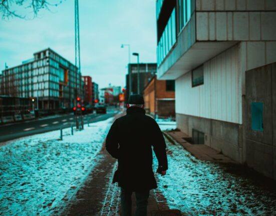 A person in warm winter attire carefully navigating a snow-covered and icy sidewalk in a city, demonstrating the need for good grip.