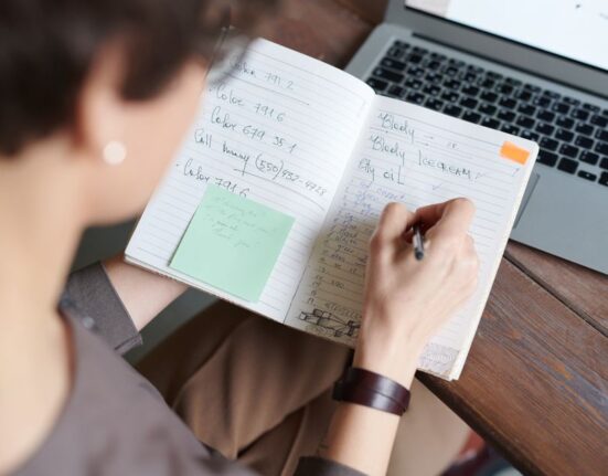 A person at a desk with a laptop open, a handwritten shopping list, and a pen, preparing for online sales.