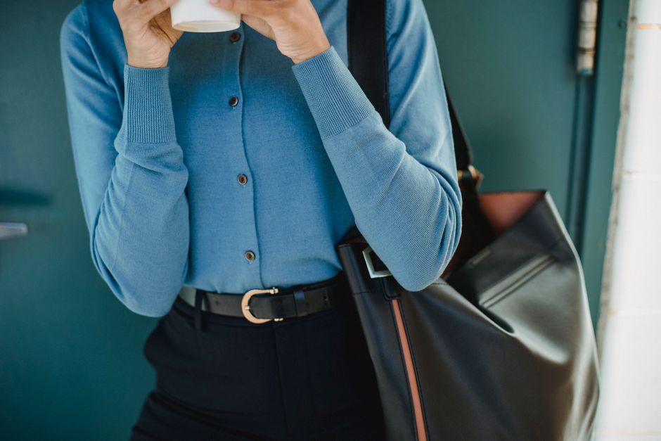 A person stylishly carrying a structured tote bag with a luggage sleeve over a rolling suitcase in a bustling airport terminal, highlighting versatility for work and travel.