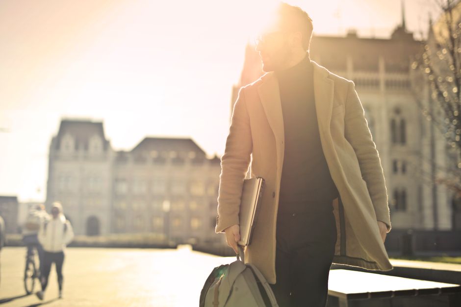 A stylish man wearing a smart casual outfit, including a grey blazer, white shirt, and dark trousers, walking through a historic European city square.