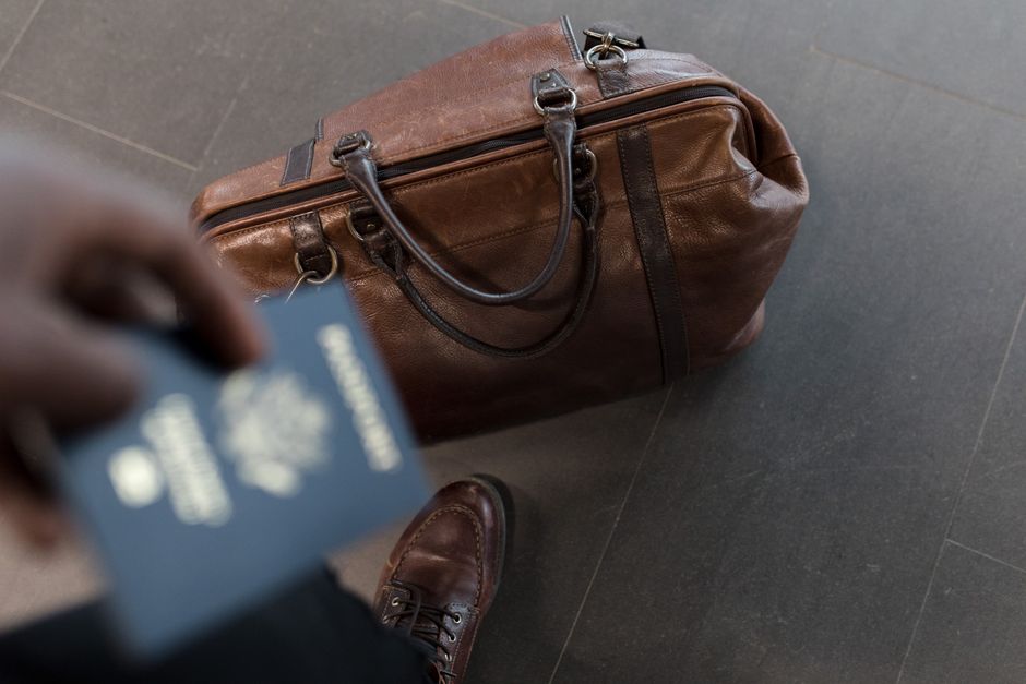 A classic, well-traveled real leather carry-on bag with a rich patina, positioned on a luggage cart in a bustling airport terminal, ready for an international flight.
