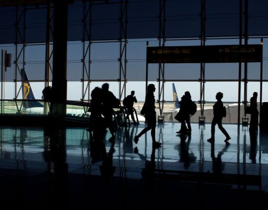 A person with a hardshell carry-on suitcase, featuring spinner wheels, walking through a brightly lit, modern airport terminal.