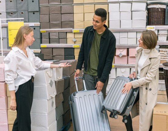 A person in a store carefully comparing a vegan leather tote bag and a real leather crossbody bag, with travel luggage visible in the background, making a decision for the best bags for travel and work.