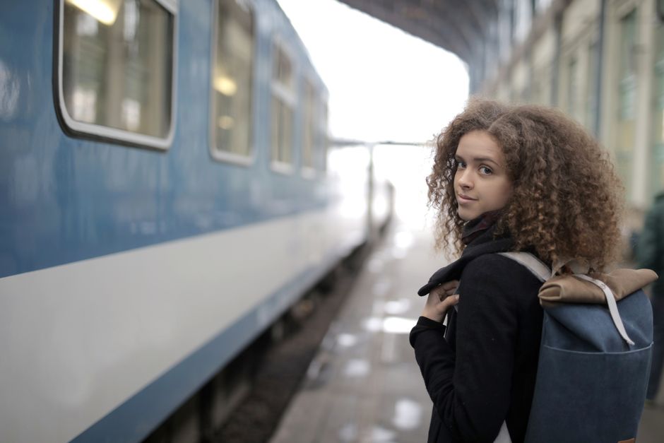 Stylish woman walking through a train station concourse, wearing wide-leg trousers, a knit top, and a light jacket, with luggage.