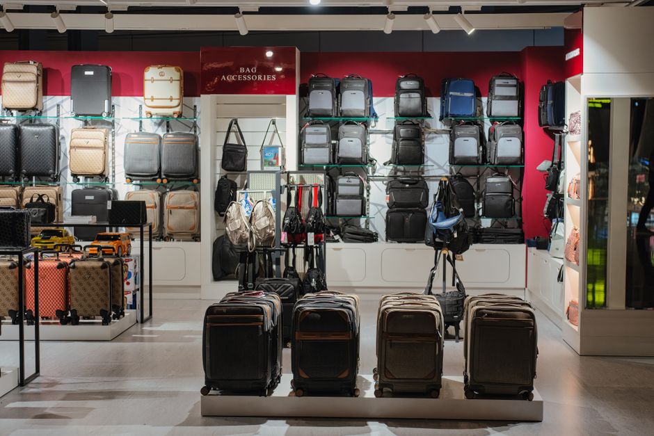 Two travelers, one with a rolling carry-on suitcase and another with a large travel backpack, standing in an airport terminal discussing their luggage choices.