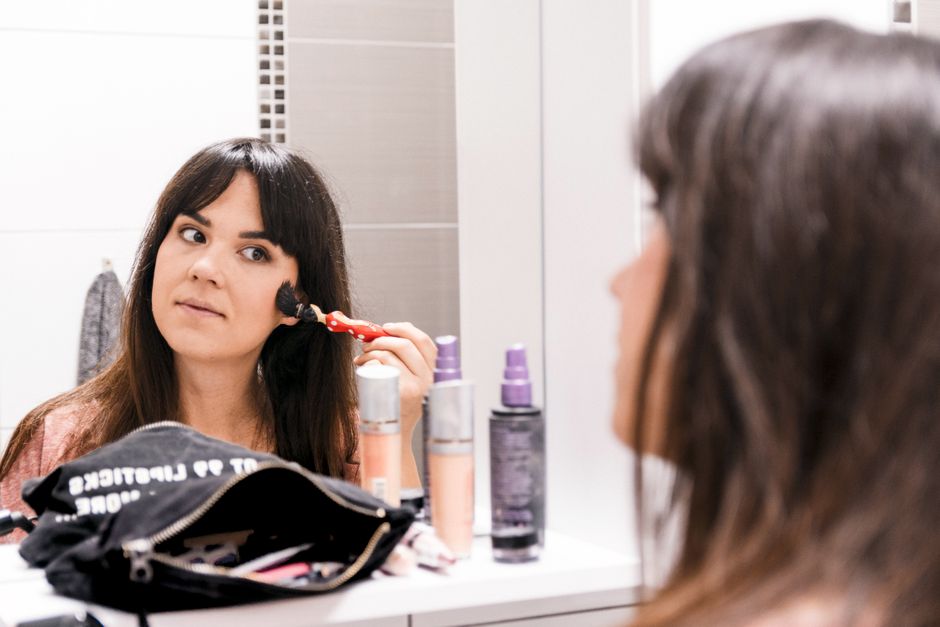 A smiling woman applying blush to her cheekbones in a handheld mirror, showing a natural makeup look.