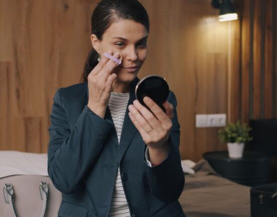 A woman in an airport lounge applying a light layer of tinted moisturizer, smiling at her compact mirror, showcasing practical beauty and skincare for travel.