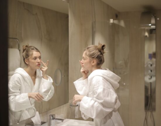 A woman in her 30s or 40s gently applying a cream to her face while looking in a bathroom mirror, representing a typical daily skincare routine.