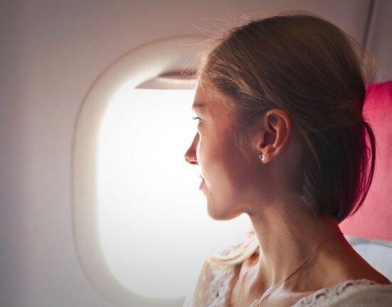 A woman wearing soft, layered clothing comfortably rests on an airplane seat, looking out the window during a long flight.