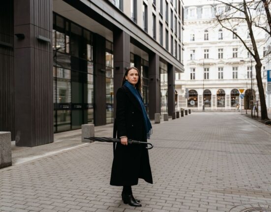 A woman in a long, elegant grey wool coat, turtleneck, and dark trousers walking confidently on a city street, demonstrating stylish women's winter outfits for cold cities.
