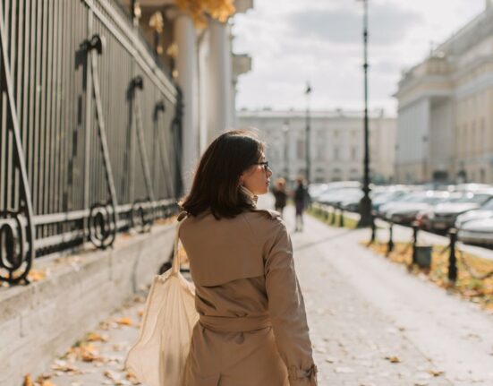 A woman in a stylish trench coat, dark jeans, and comfortable sneakers walking down a charming European city street, perhaps in Paris or London.