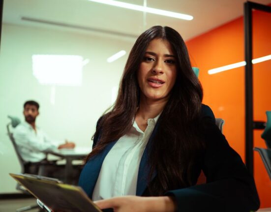 A professional woman smiles confidently in a bright, modern hybrid office space, wearing a cream blazer, dark tailored trousers, and a simple blouse, epitomizing easy workwear outfits for hybrid offices.
