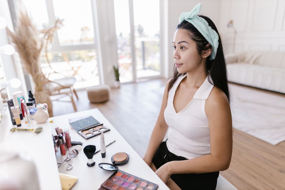 A person's hands delicately layering a serum onto their face, with an array of skincare bottles visible on a minimalist vanity in a sunlit urban apartment.