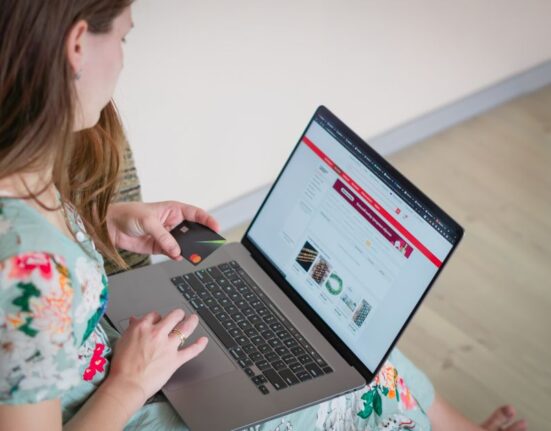 A woman sits at a table, happily shopping online on her laptop, surrounded by credit cards and a coffee cup, illustrating savvy deal hunting.