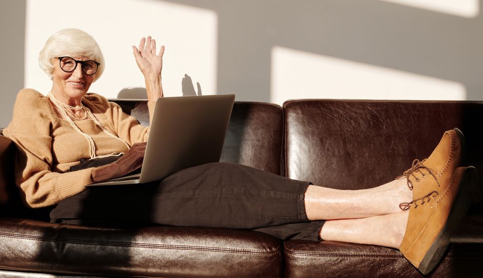 A woman smiling and pointing at a laptop screen displaying an online shoe store's product page, with a pair of shoes visible on the screen and her feet resting comfortably nearby.